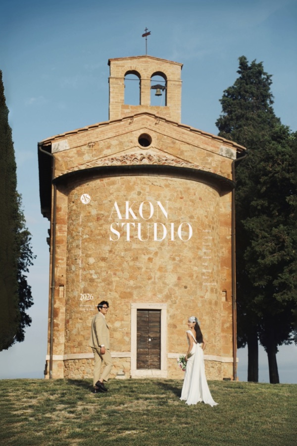 Couple on a lawn in front of a small Romanesque stone chapel in Tuscany, bride in a white gown with a floral headpiece, groom in a beige suit, tall cypresses and blue sky behind