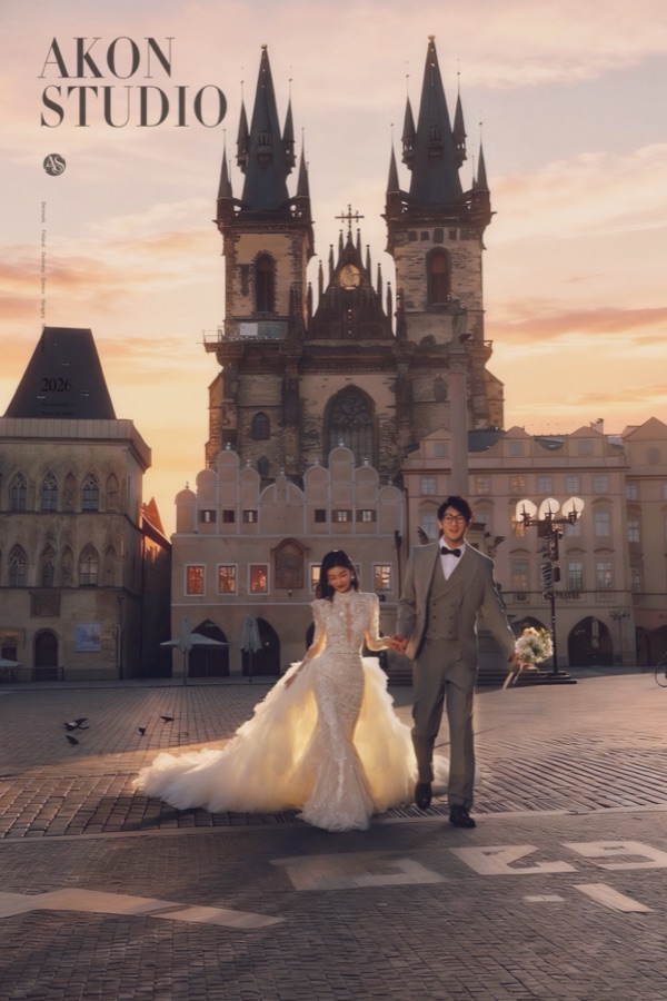 Couple walking across the cobbles of Prague Old Town Square at dusk, bride in a beaded gown with a long tulle train, groom in a grey-green suit, Tyn Church towers behind them in pink light