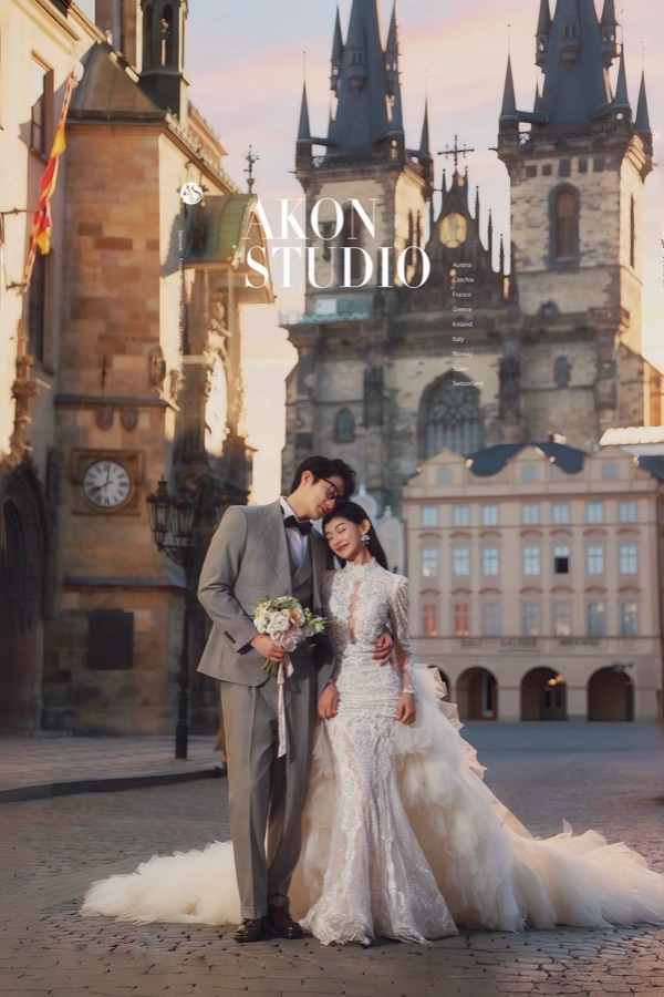 Couple in a lace gown and grey suit in an empty Old Town Square at sunrise, pink sky behind the twin spires of Tyn Church