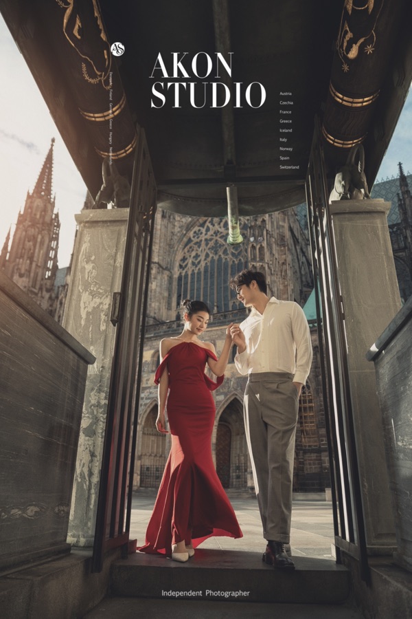 Bride in a true-red mermaid gown and groom in a white shirt and grey trousers holding hands under a gothic stone portal in Prague