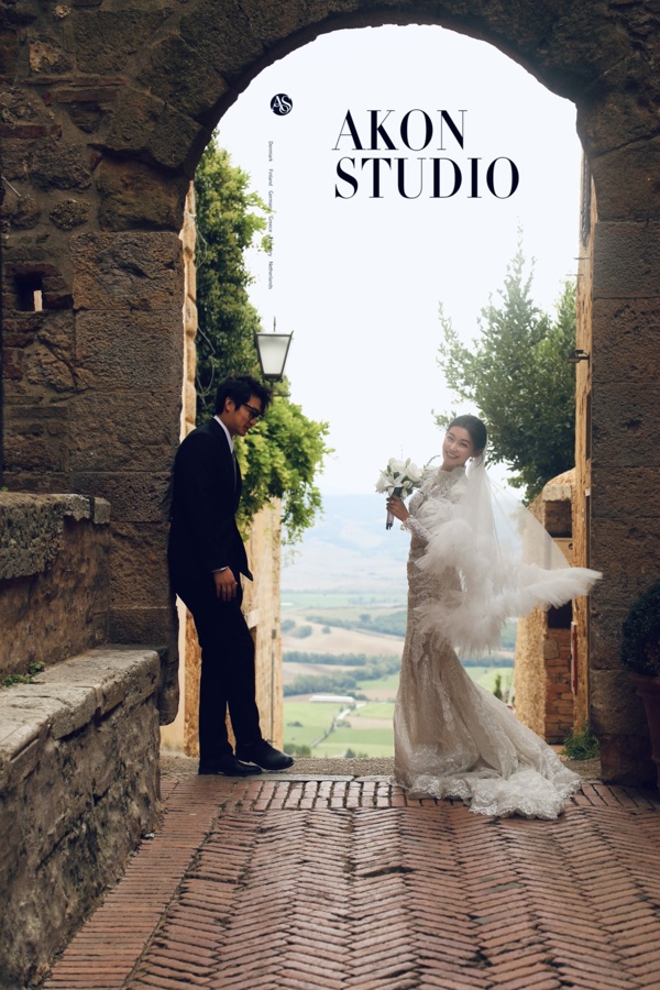 Bride in a lace gown with a feather veil holding white flowers under a Tuscan stone arch, groom in a black suit waiting by the wall