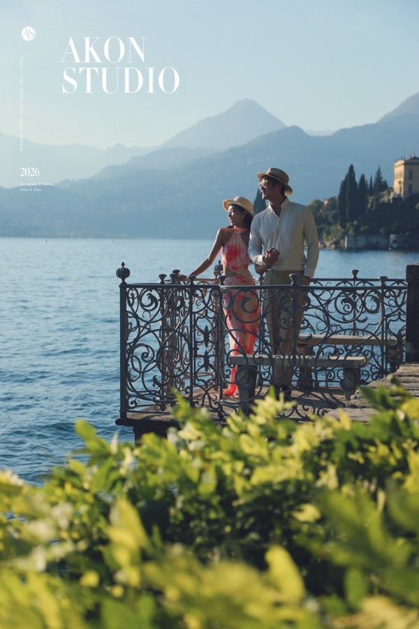 Couple in holiday wear on a wrought-iron balcony over Lake Como, bride in a pink slip dress and straw hat, groom in a cream shirt