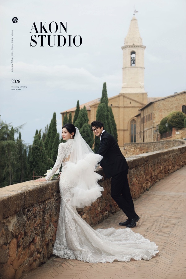 Bride's long veil lifting in the wind on Pienza's stone wall, cypress trees and a church bell tower behind